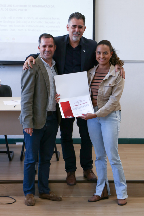 Três pessoas posam sorrindo para uma foto. A mulher à direita segura um certificado com a marca da Ufop. Os dois homens a seu lado estão sorrindo, com braços sobre os ombros dela.