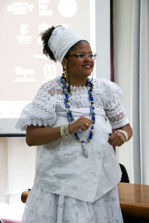 Mulher negra sorridente, vestida com roupa tradicional branca de matriz africana, com turbante, colares e pulseiras. Ela está em pé, em ambiente interno, com um projetor ao fundo exibindo logotipos.