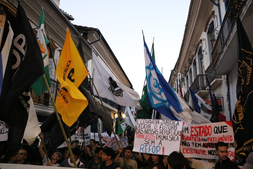 Manifestantes marcham por uma rua estreita do centro histórico, segurando bandeiras e cartazes de protesto. Há várias bandeiras pretas, amarelas, verdes e azuis erguidas acima da multidão. Alguns cartazes escritos em português criticam decisões políticas 