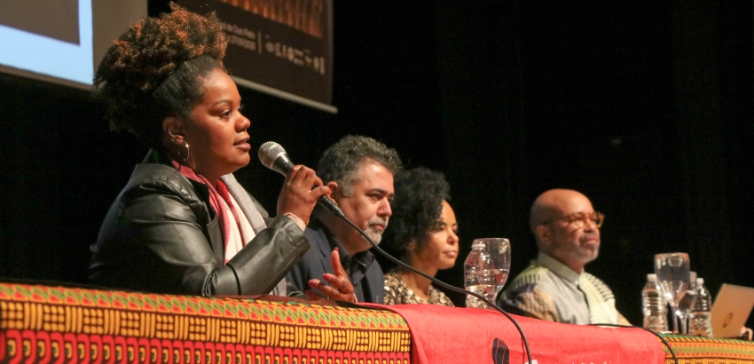Imagem de um painel com quatro pessoas. À esquerda, mulher negra de cabelo volumoso e roupas escuras, fala ao microfone. Ao centro, homem com cabelo grisalho e camisa escura. À direita, mulher negra com cabelo curto e homem negro com camisa clara.