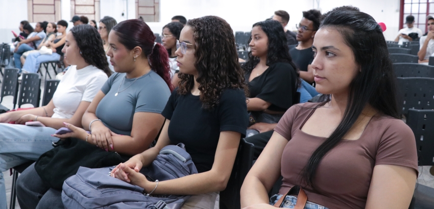 Grupo de jovens sentados em cadeiras de um auditório, assistindo atentamente a uma apresentação. A maioria está com expressão séria e concentrada.