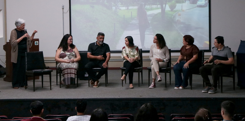Sete pessoas estão sentadas em cadeiras no palco de um auditório, participando de uma conversa, enquanto uma mulher em pé, à esquerda, fala ao microfone. Ao fundo, há uma projeção com imagem da UFOP.