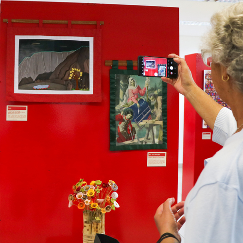 mulher de cabelo grisalho e blusa clara fotografa uma tapeçaria que representa uma cena religiosa com seu smartphone, em frente a uma parede vermelha. Ao redor, há outra tapeçaria com paisagem escura e um arranjo de flores de biscuit em um vaso de bambu