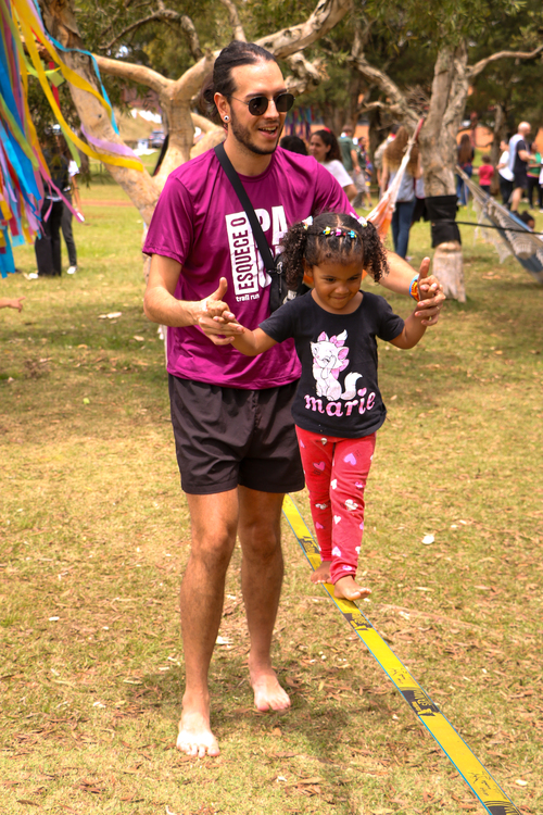 Um homem barbudo de óculos de sol, camiseta magenta e shorts pretos, descalço, sorri enquanto segura as mãos de uma menina pequena, ajudando-a a caminhar descalça sobre uma fita de slackline amarela em um parque.