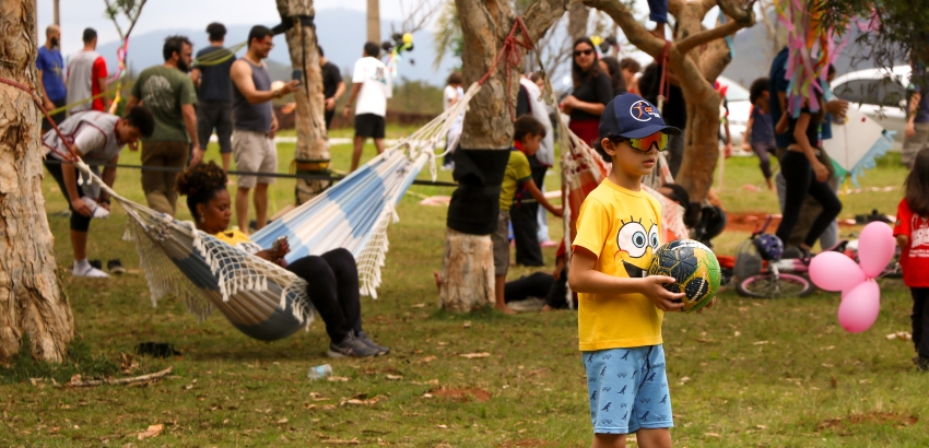 Um menino de óculos de sol, boné azul e camiseta amarela segura uma bola de futebol em um parque gramado, rodeado por árvores. Ao fundo, diversas pessoas interagem, e uma mulher relaxa em uma rede listrada.
