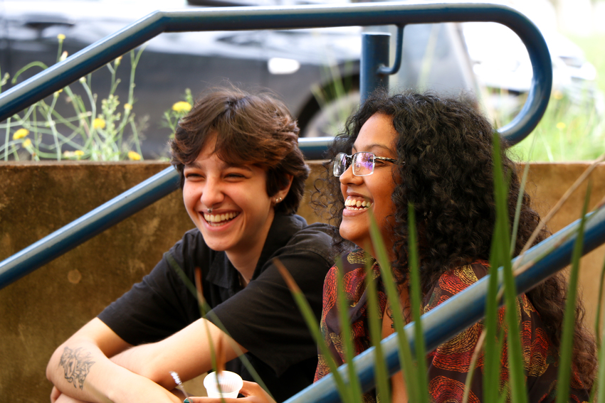 Duas jovens sorridentes, uma com cabelo curto e óculos escuros e outra de cabelo cacheado e óculos de grau, sentam-se em degraus, entre hastes de grama e um corrimão azul