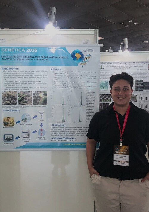 Pessoa sorri em frente a um pôster científico exposto em congresso, que apresenta pesquisa sobre genética de cactos. Usa crachá de identificação do evento e está em ambiente de exposição acadêmica.