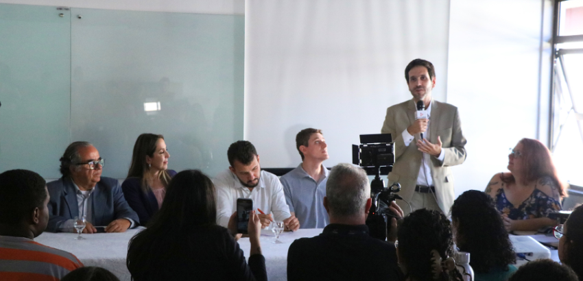 Foto da mesa redonda compondo seis integrantes, cinco deles sentados ouvindo o assessor Otávio Penna Pieranti, a foto também contém a plateia prestando atenção ao pronunciamento no canto inferior