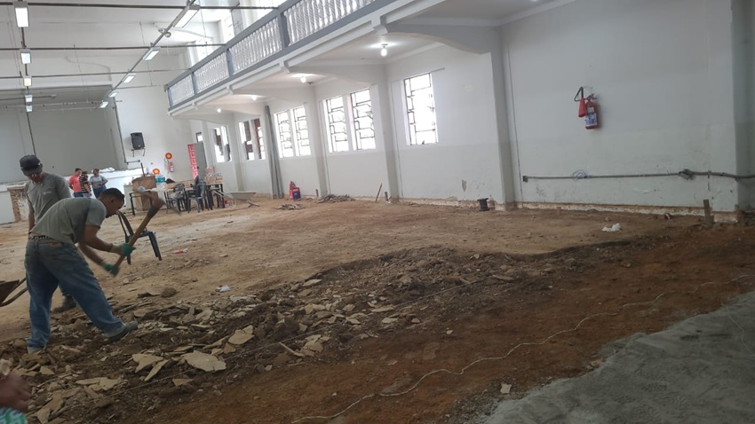 Trabalhadores realizam obra em grande salão interno, retirando o piso antigo e expondo o chão de terra. O local tem janelas amplas, mezanino com guarda-corpo branco e iluminação no teto. Ferramentas e entulhos estão espalhados pelo chão.