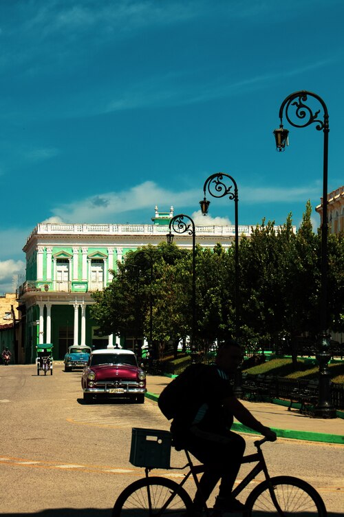 Silhueta de ciclista pedalando em rua ensolarada, com postes ornamentais e prédios coloniais verde-claros ao fundo; carros antigos estacionados e céu azul intenso completam a cena urbana.