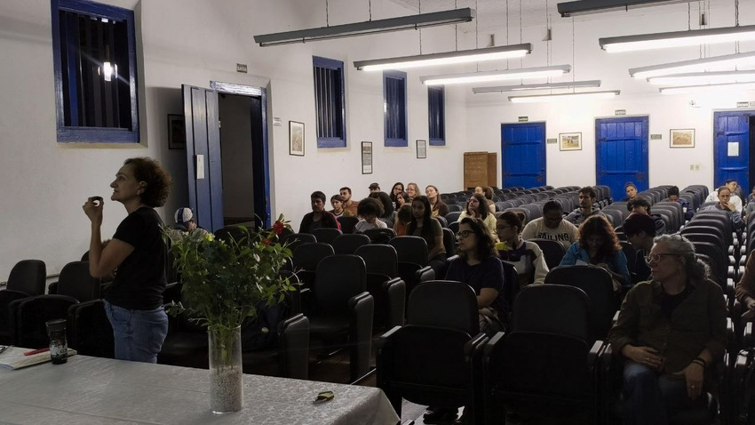 imagem mostra a professora Regina Horta da Ufop , ela está em pé e faz uma pergunta ao professor Manoel. Os demais estão sentados acompanhando o diálogo.