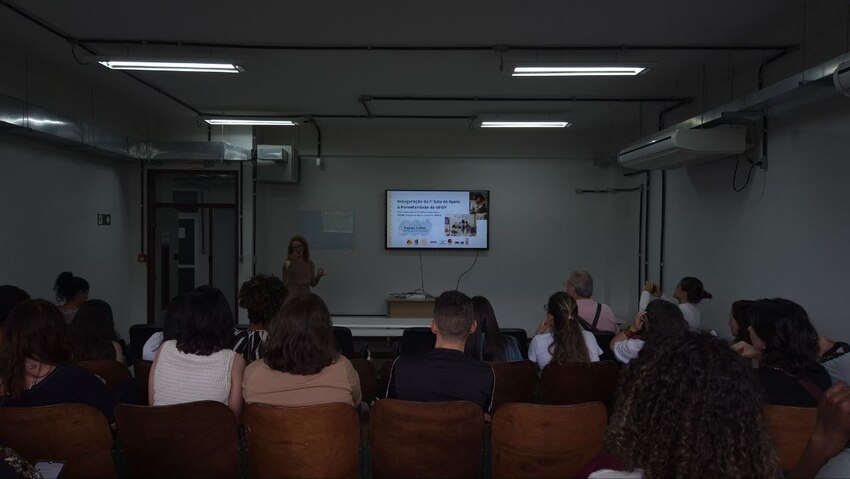 Público sentado assiste a apresentação em sala de aula escura, com tela ao fundo exibindo slide sobre ações de saúde. Uma mulher, em pé, fala perto da TV enquanto participantes observam atentamente no ambiente iluminado por luzes artificiais.
