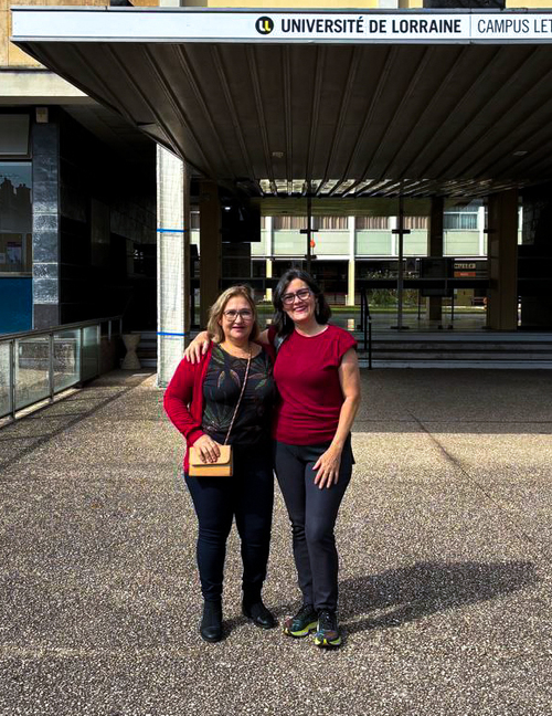 Duas mulheres sorrindo em área externa, diante de um prédio com colunas e janelas, usando roupas em tons de vermelho e preto.