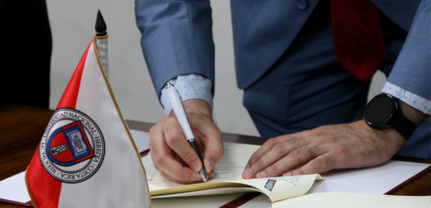 Pessoa em traje formal assina documento em livro de registros durante cerimônia institucional. Ao lado, pequena bandeira da Universidad Nacional da Costa Rica sobre a mesa, simbolizando acordo de cooperação internacional entre universidades.