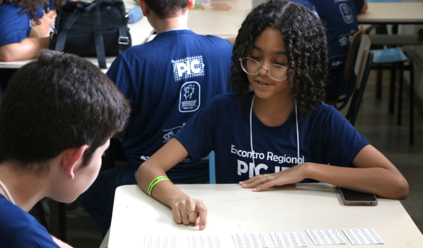 Estudante com camiseta azul do Encontro Regional PIC OBMEP participa de atividade em grupo, apontando para uma folha com números impressos sobre a mesa. Ao seu lado, outro participante acompanha atentamente a tarefa.