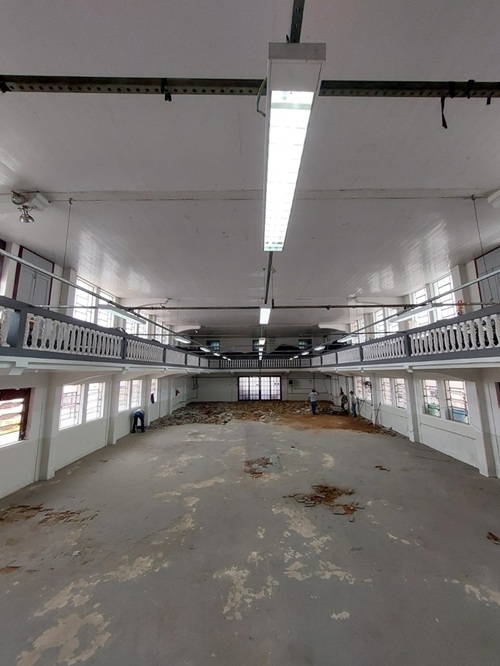 Imagem de um grande salão interno em reforma, com piso desgastado e parcialmente removido. Há andaimes, entulho no chão e alguns trabalhadores ao fundo. O espaço possui mezaninos laterais com guarda-corpos brancos e luminárias no teto.          Perguntar 