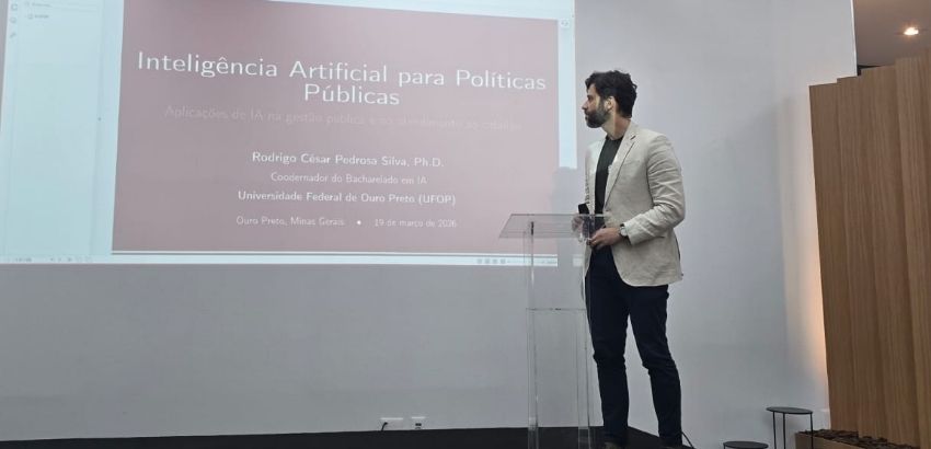 Um homem de cabelo e barba preta, usando uma calça preta e um blazer bege, apresenta uma palestra em pé em uma sala de paredes brancas. Um slide está projetado na parede com o título “Inteligência Artificial para Políticas Públicas”.