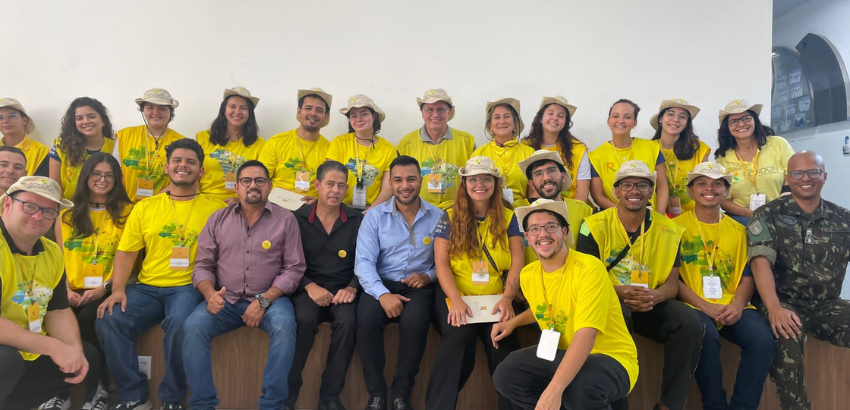 Fotos de vinte e cinco pessoas enfileiradas em duas fileiras, sorrindo de frente para a câmera, a maioria das pessoas usando a blusa amarela do projeto enquanto outras estão de farda