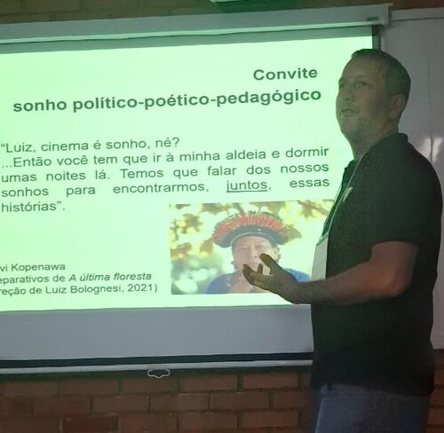 Homem em pé diante de um slide projetado em tela, que exibe o título “Convite: sonho político-poético-pedagógico” e uma citação de Davi Kopenawa sobre cinema e sonhos, com uma imagem de um homem indígena usando cocar colorido.