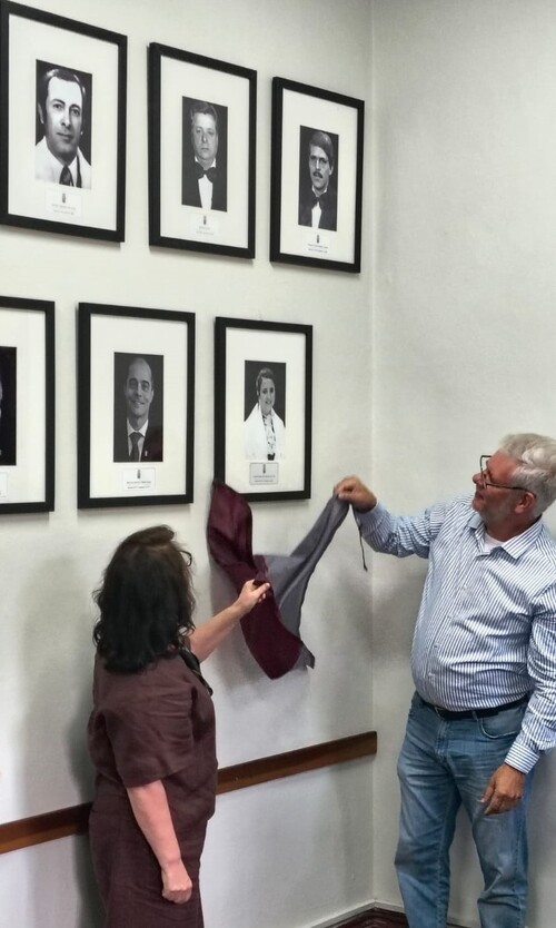 Um homem e uma mulher realizam o descerramento de um quadro em uma parede com retratos emoldurados. Eles puxam um tecido bicolor que cobria a nova fotografia enquanto observam o resultado da cerimônia