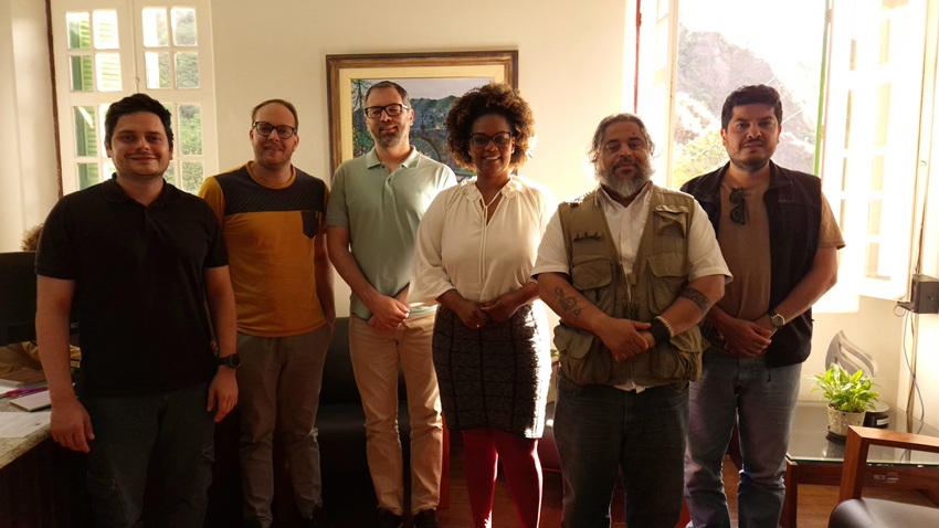 Seis pessoas posam sorrindo em ambiente interno iluminado pela luz natural vinda das janelas. Ao fundo, há um quadro com paisagem e uma planta próxima à janela. O grupo está em clima formal, lado a lado, olhando para a câmera.