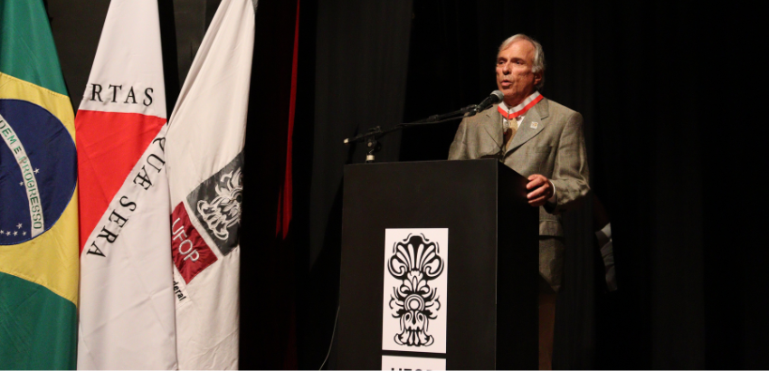 O professor Rinaldo, um homem de aproximadamente 70 anos, tem pele e cabelos brancos e lisos. Usa um terno cinza e fala ao microfone.