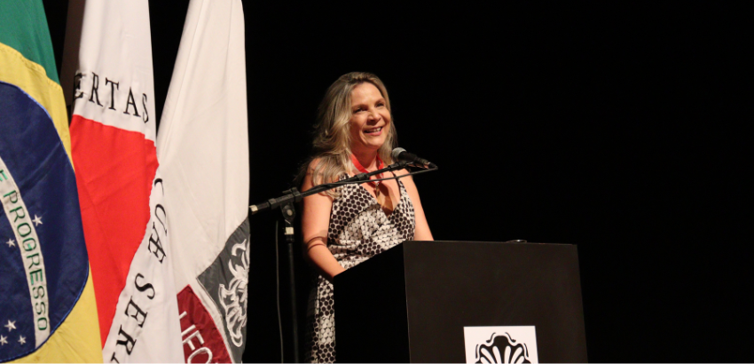 Roneimar é loira, usa um vestido preto e branco e sorri. Ela está no púlpito que tem a marca da UFOP.