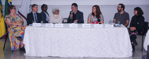 Pessoas sentadas atrás de uma mesa com toalha branca participam de uma mesa-redonda ou conferência. Um dos participantes fala ao microfone, enquanto os demais escutam atentamente. Há placas de identificação sobre a mesa.