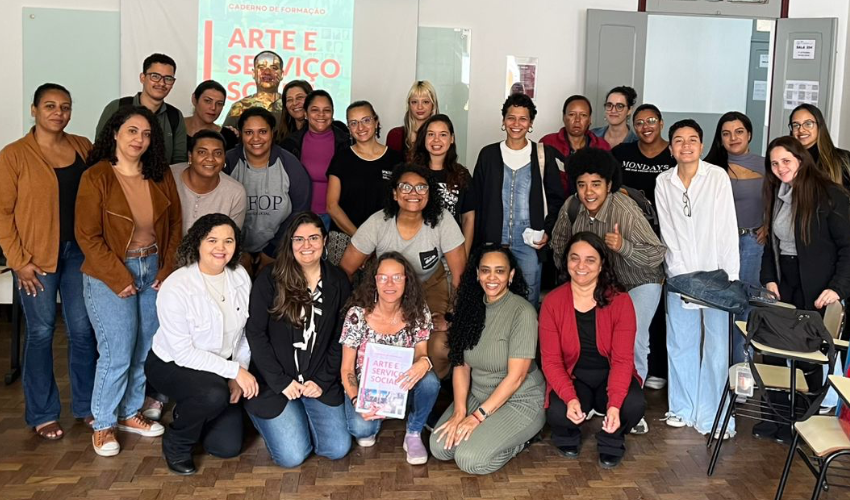 Grupo de cerca de 25 pessoas posa sorridente em sala de aula, com carteiras, mochilas e lanches ao fundo. No quadro, projeta-se a capa do “Caderno de Formação: Arte e Serviço Social”. Uma participante segura o material impresso na frente.