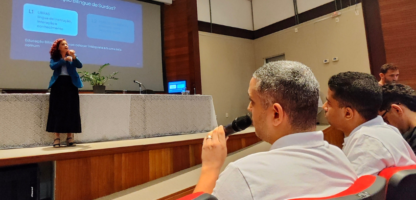 Palestrante apresenta sobre educação bilíngue de surdos em um auditório. No palco, a tela exibe “O que é Educação Bilíngue de Surdos?”. Em primeiro plano, um tradutor de Libras, de camisa branca, segura um microfone.