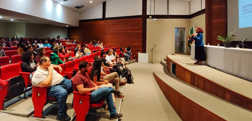 Auditório com público sentado em cadeiras vermelhas enquanto uma palestrante fala no palco, ao lado de um telão com apresentação projetada.