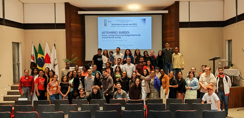Foto de grupo em auditório, com dezenas de pessoas reunidas diante de um telão com a apresentação “Setembro Surdo: lutas, conquistas e protagonismo do movimento surdo”. Bandeiras aparecem ao lado do palco.