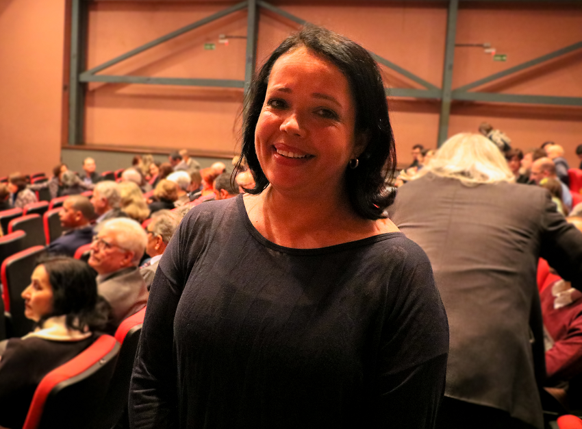 Foto de Sílvia, uma mulher negra de cabelos pretos na altura dos ombros, sorrindo. Atrás dela, está o auditório do Centro de Convenções.