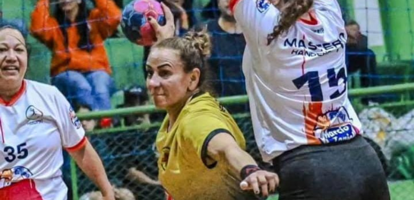 Jogadora de handebol em uniforme amarelo arremessa a bola com expressão de foco e força; ao lado, atletas em branco acompanham a jogada. Público desfocado assiste ao fundo da quadra.