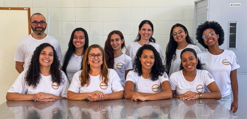 Imagem mostra dez pessoas, composto por nove mulheres e um homens, em uma sala com paredes de azulejos brancos.Todos usando camisetas brancas com o logotipo “NEPAN” no peito. Na frente, quatro mulheres estão sentadas e os demais em pé, e visível uma mesa.