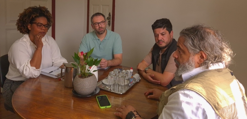 Quatro pessoas estão sentadas ao redor de uma mesa de madeira em uma reunião. Há cadernos, copos, uma planta, celular e garrafa sobre a mesa. Todos parecem concentrados na conversa.