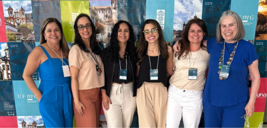 Seis mulheres posam sorridentes lado a lado em frente a um painel colorido com imagens e logotipos de universidades mineiras. Elas usam crachás de identificação, transmitindo clima de evento acadêmico e de confraternização.