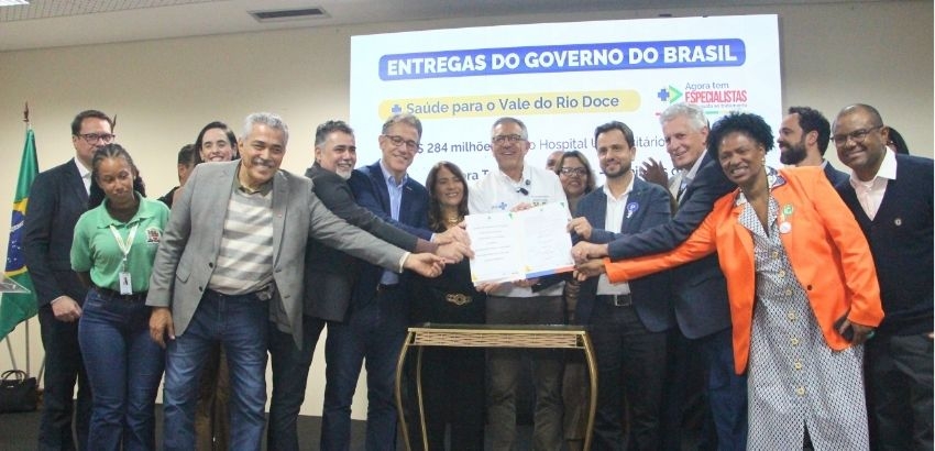 Fotografia de uma cerimônia institucional realizada em ambiente interno. Cerca de treze pessoas estão reunidas em frente a uma tela de projeção que exibe o título “Entregas do Governo do Brasil” e informações sobre investimentos em saúde para o Vale do Ri