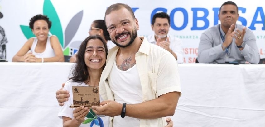 Estudante sorri enquanto recebe uma placa de Menção Honrosa durante cerimônia do IX Congresso Baiano de Engenharia Sanitária e Ambiental (COBESA). Ao seu lado, o professor orientador também sorri para a câmera. Ao fundo, integrantes da mesa do evento acom