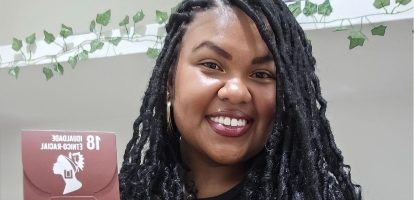 Mulher negra sorrindo para a câmera, com cabelos longos trançados e brincos dourados, segurando uma placa com o texto “18º Encontro-Cacional” e um símbolo de perfil feminino. Ao fundo, há uma parede clara decorada com folhas verdes.