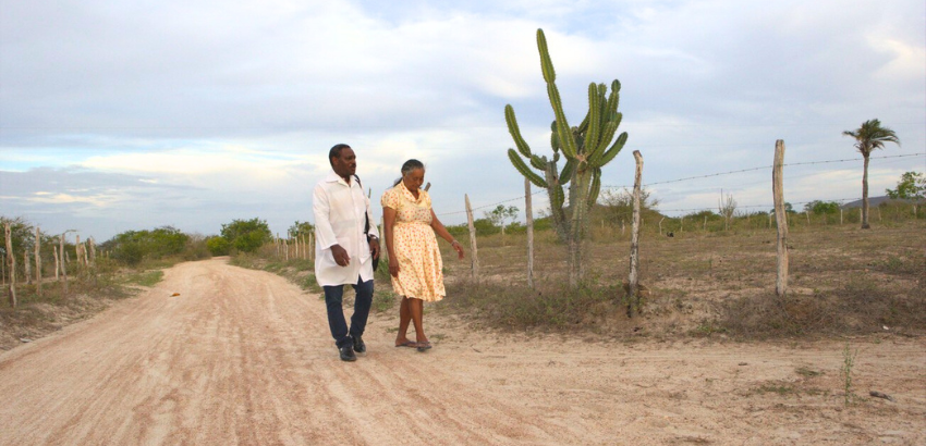 Um homem com um jaleco branco e uma mulher de vestido amarelo e detalhes vermelhos ambos negros caminham lado a lado por uma estrada de terra