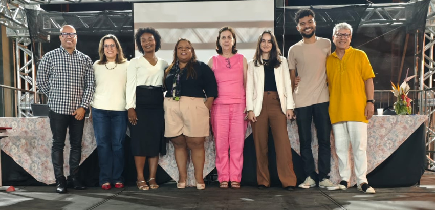 Oito pessoas estão lado a lado em um palco, posando para uma foto em grupo. Atrás delas há uma tela e estruturas metálicas. Todos sorriem e usam roupas casuais e coloridas.