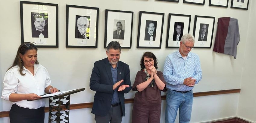 Quatro pessoas participam de uma cerimônia em uma sala com retratos na parede. À esquerda, uma mulher lê em um púlpito; ao centro, um homem e uma mulher aplaudem; à direita, um homem observa. Um dos quadros está coberto por tecido