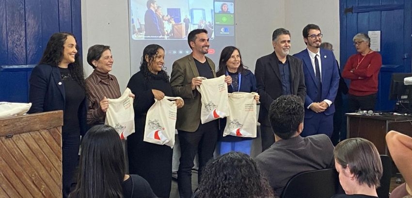 Em uma sala de eventos, sete pessoas estão alinhadas na frente de um público sentado, sorrindo para a foto. Quatro delas seguram sacolas brancas de um evento institucional. Ao fundo, uma tela de projeção exibe uma videoconferência com participantes remoto