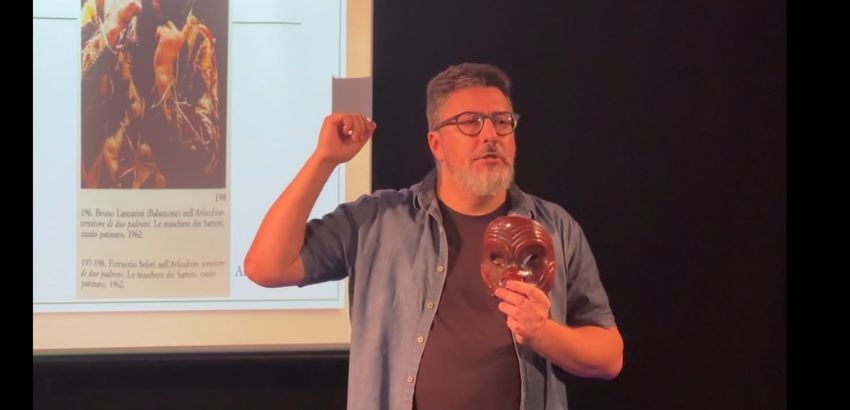 Homem de óculos, barba grisalha e camisa azul aberta sobre camiseta escura faz uma apresentação em um palco. Ele segura uma máscara marrom esculpida em uma das mãos e gesticula com a outra enquanto fala. Ao fundo, há uma tela de projeção ex