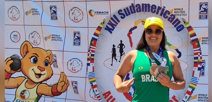Imagen de Marinalva com medalhas após conquistar vice-campeonato Atletismo Master. A atleta usa um óculos esculo, camisa verde escrita "BRASIL" em amarelo e usa boné. Ao fundo, observa-se o mascote e um painel com os patrocinadores.