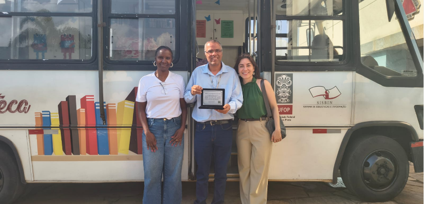 A imagem mostra três pessoas em pé, sorrindo, em frente a um ônibus do projeto Carro Biblioteca da UFOP.