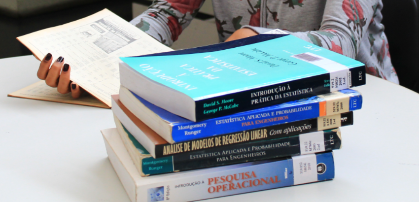 A imagem mostra uma mão segurando um caderno enquanto livros sobre estatística e pesquisa operacional estão empilhados sobre uma mesa. Os livros possuem títulos em português e estão organizados de forma ordenada.