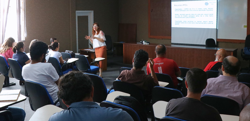 A pró-reitora de Pesquisa, Pós-Graduação e Inovação apresenta slides para docentes sentados em cadeiras com apoio. A apresentação acontece em auditório da UFOP, com projeção na parede e participantes atentos a explicação.