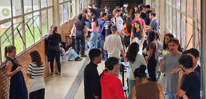 Corredor universitário com paredes de tijolo e janelas amplas, cheio de estudantes conversando em grupos.